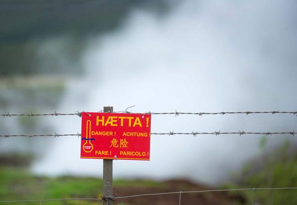 Zīme ar uzrakstu "Briesmas!" islandiešu un citās valodās brīdina tūristus par bīstamiem karstajiem avotiem aiz žoga. Islande, 20.05.2019.