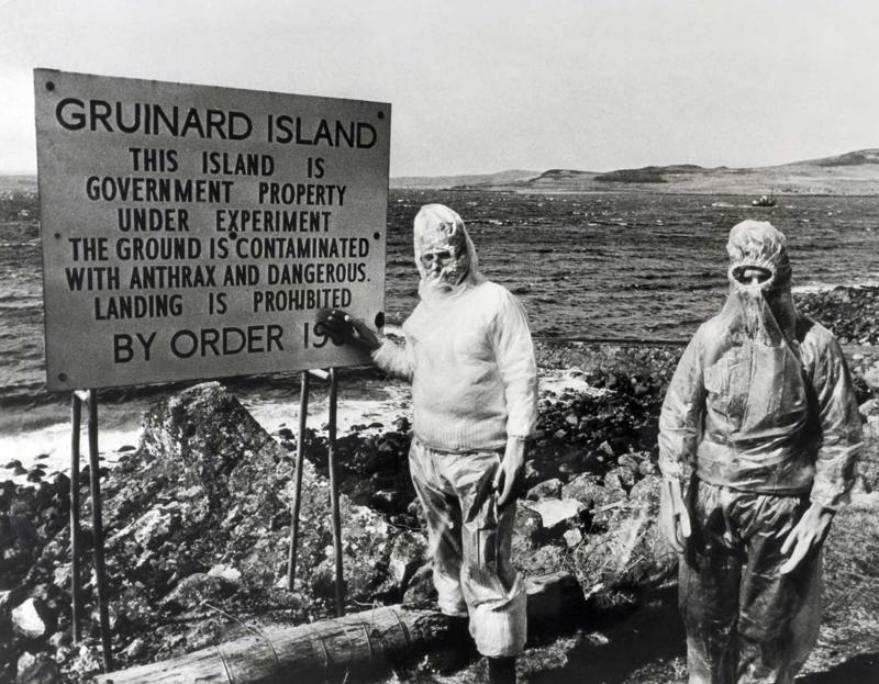 Divi vīrieši aizsargdrēbēs pie brīdinājuma zīmes par Lielbritānijas valdības bioloģisko ieroču aģenta Anthrax (Sibīrijas mēris) izmēģinājumiem Grināra salā Skotijas piekrastē.