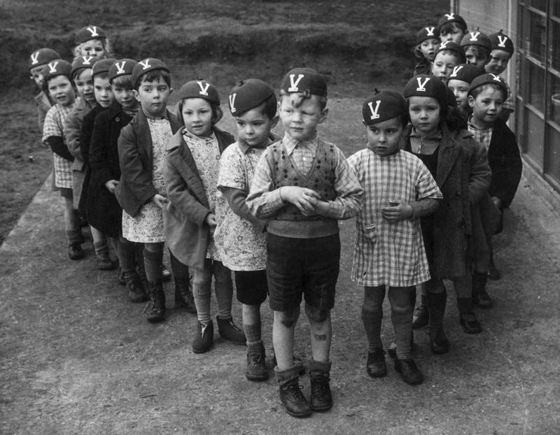Bērni nostājušies burta "V" formā ar cepurītēm, kas rotātas ar uzvaras "Victory V" zīmi. Cepurītes ziedojusi Lielbritānijas Kara palīdzības biedrība no Amerikas (British War Relief Society of America). 11.12.1941. "V for Victory" bija veiksmīga Otrā pasaules kara sabiedroto propagandas kampaņa.