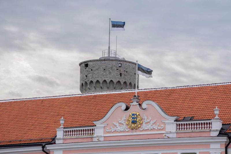 Priekšplānā Igaunijas parlamenta ēka ar Igaunijas valsts ģerboni, aiz tās Igaunijas valsts karogs Tallinas Garā Hermaņa tornī. 31.08.2017.