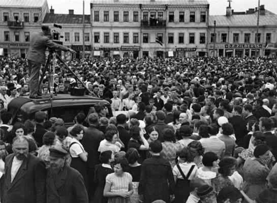 Daugavpils iedzīvotāji Tirgus laukumā sagaida Latvijas Ministru prezidentu Kārli Ulmani un valdības locekļus vizītes laikā Latgalē, priekšplānā uz automašīnas jumta visticamāk fotogrāfs un kinooperators Eduards Kraucs ar kameru. Daugavpils, 08.1934.