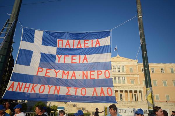 Grieķijas karogs ar uzrakstu grieķu valodā "Izglītība, veselības aprūpe; ūdens un elektrība ir bezmaksas visiem cilvēkiem" pie Grieķijas parlamenta ēkas protestu laikā. Atēnas, 09.07.2014.