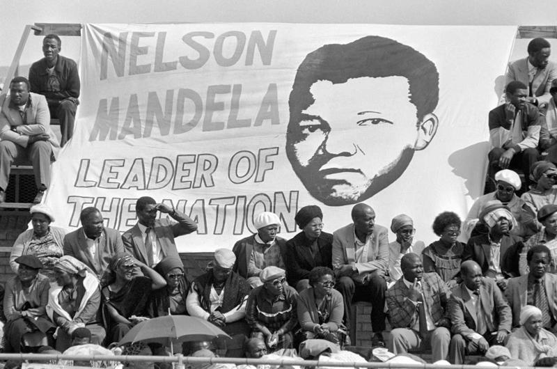 Dienvidafrikāņi tur baneri ar Nelsona Mandelas portretu un saukli “Nelsons Mandela, nācijas līderis” deviņu Guguletu nemieros nogalināto cilvēku bērēs. Keiptaunas apkārtne, Dienvidāfrika. 07.09.1985. 